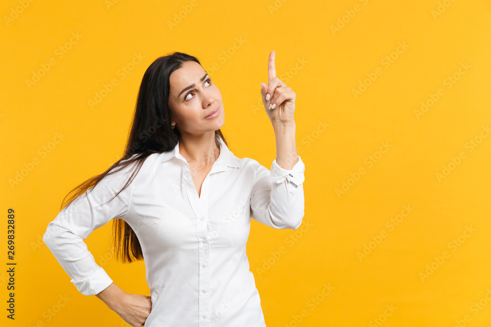 Portrait of puzzled pensive young woman in white casual shirt looking and pointing index finger up isolated on yellow orange wall background in studio. People lifestyle concept. Mock up copy space. Portrait of puzzled pensive young woman in white casual shirt looking and pointing index finger up isolated on yellow orange wall background in studio. People lifestyle concept. Mock up copy space.