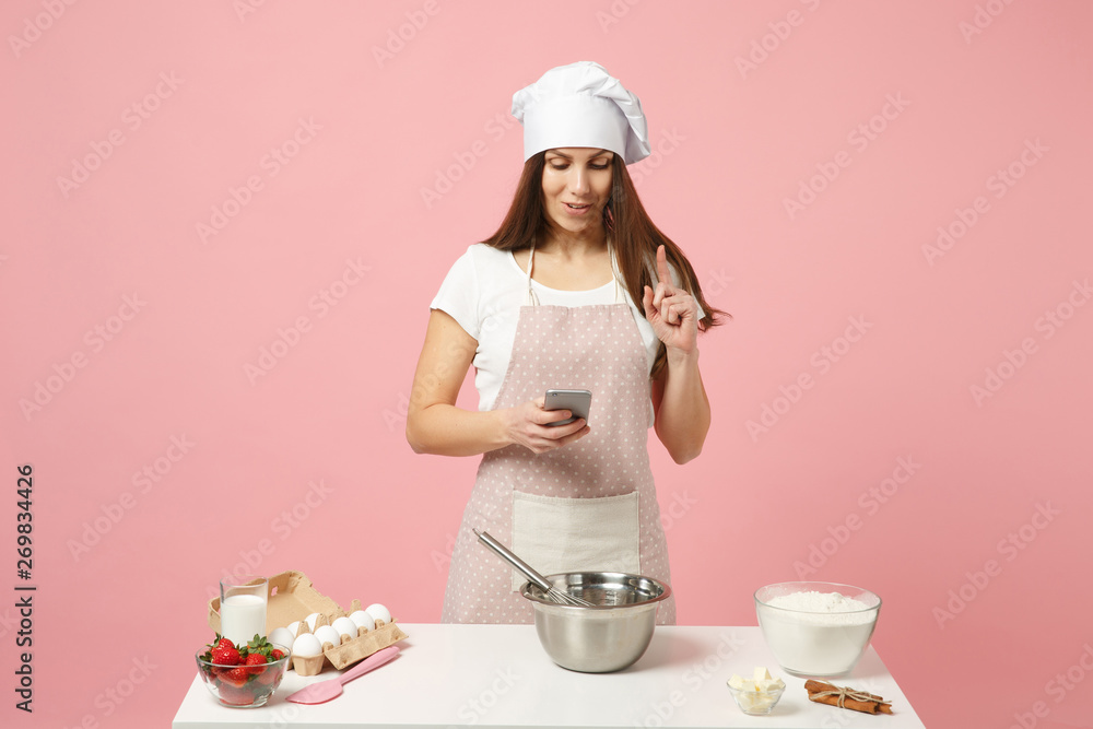 Chef cook confectioner or baker in white t-shirt, toque chefs hat cooking cake or cupcake at table hold using mobile phone isolated on pink pastel background in studio. Mock up copy space food concept