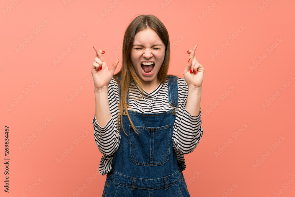 Young woman with overalls over pink wall with fingers crossing and wishing the best Young woman with overalls over pink wall with fingers crossing and wishing the best