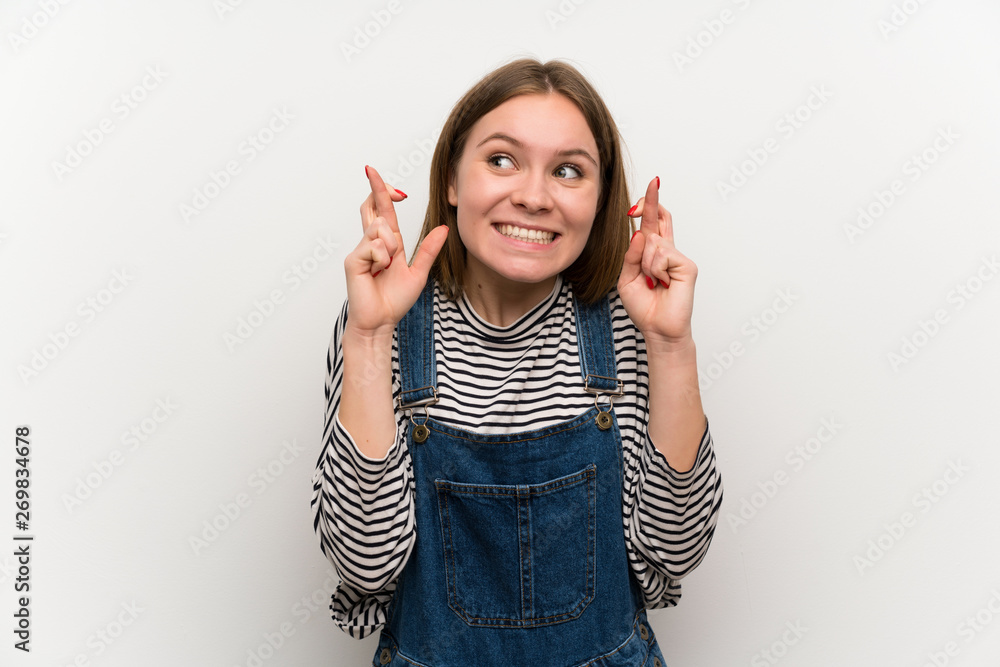 Young woman in dungarees over white wall with fingers crossing