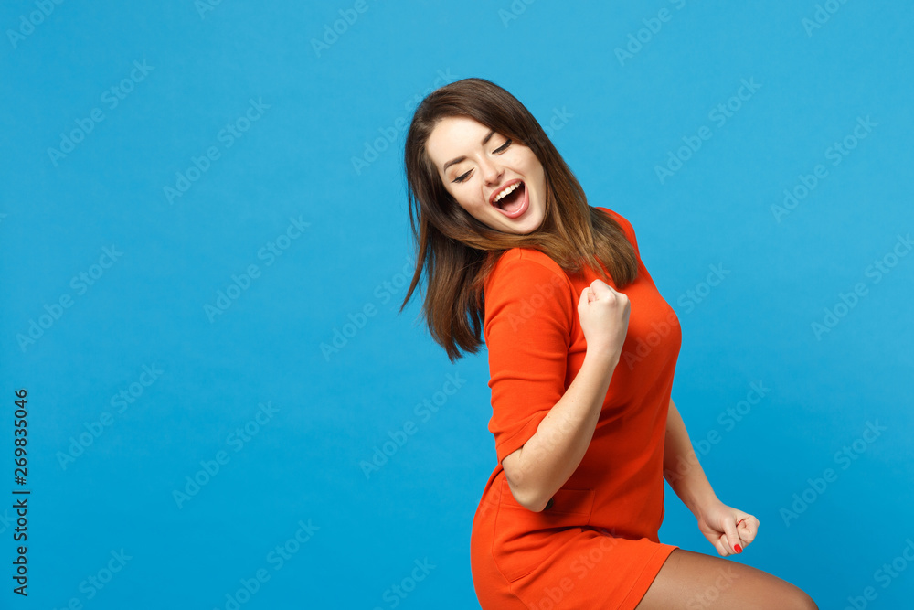 Beautiful excited brunette young woman wearing red orange dress doing winner gesture, say Yes isolated over blue wall background, studio portrait. People lifestyle fashion concept. Mock up copy space. - obrazy, fototapety, plakaty 
