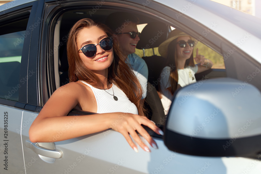 Group of friends rented a car on summer road trip and arrived to the sea beach. Woman in glasses looks out of the car window. Passanger girl having fun with friends in vehicle. Travel lifestyle.