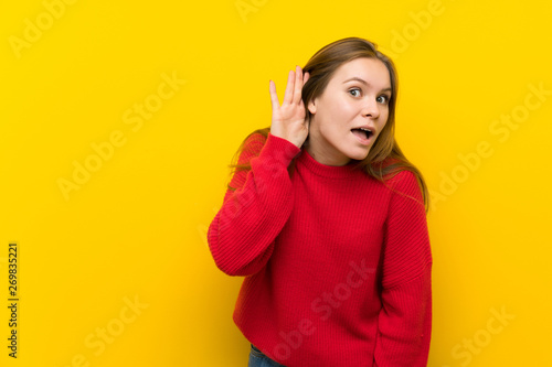 Young woman over yellow wall listening to something by putting hand on the ear © luismolinero