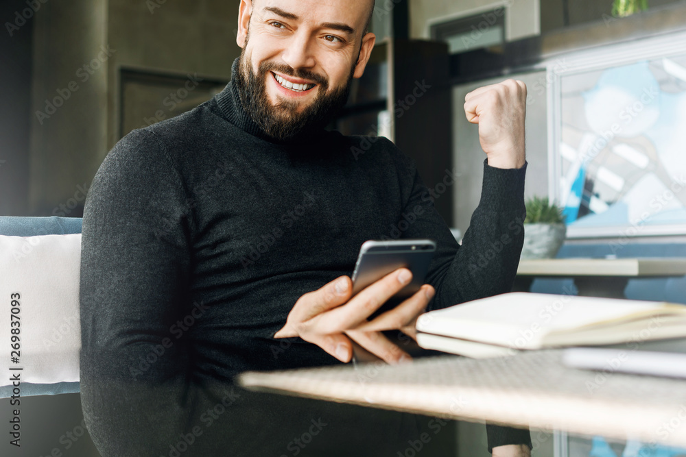 Bearded middle-aged man holds smartphone and is emotionally happy about winning, winning while sitting in cafe at table. Bearded middle-aged man holds smartphone and is emotionally happy about winning, winning while sitting in cafe at table.