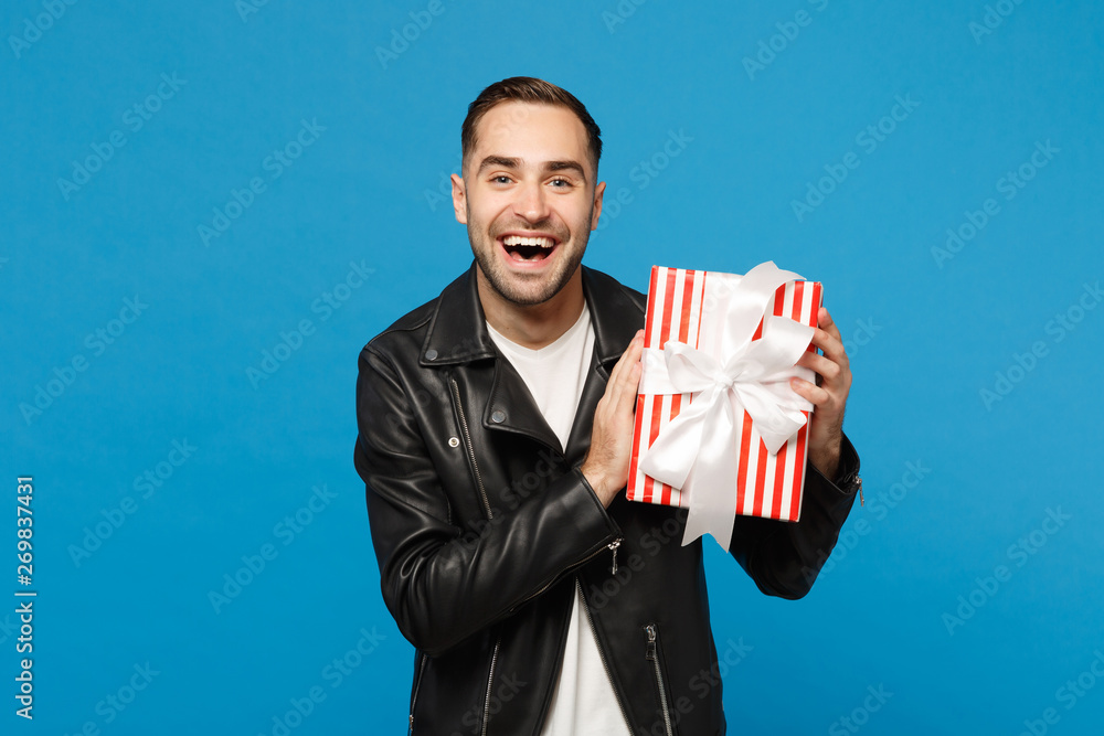Handsome stylish young bearded man in black leather jacket white t-shirt hold gift box isolated on blue wall background studio portrait. People sincere emotions lifestyle concept. Mock up copy space. Handsome stylish young bearded man in black leather jacket white t-shirt hold gift box isolated on blue wall background studio portrait. People sincere emotions lifestyle concept. Mock up copy space.