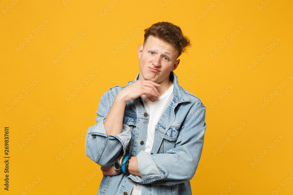 Displeased young man in denim casual clothes looking camera, put hand prop up on chin isolated on yellow orange background in studio. People sincere emotions, lifestyle concept. Mock up copy space. Displeased young man in denim casual clothes looking camera, put hand prop up on chin isolated on yellow orange background in studio. People sincere emotions, lifestyle concept. Mock up copy space.