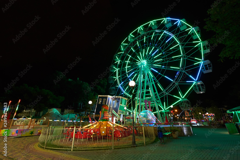 Amusement park at night with a ferris wheel and carousels. Glowing ferris wheel