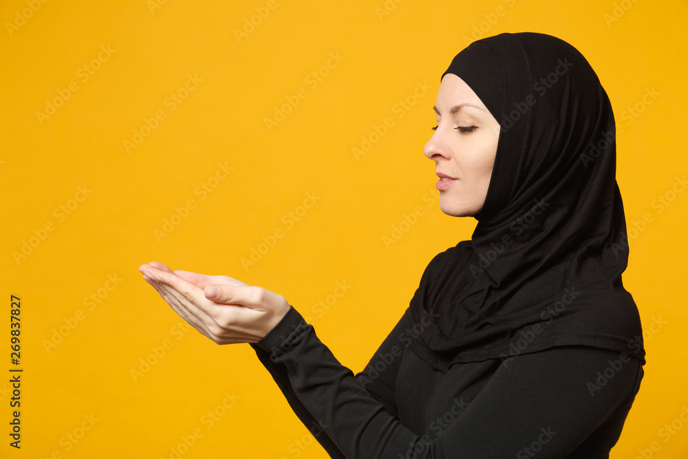 Side view young arabian muslim woman in hijab black clothes hold in hands empty workspace isolated on yellow wall background, studio portrait. People religious lifestyle concept. Mock up copy space.