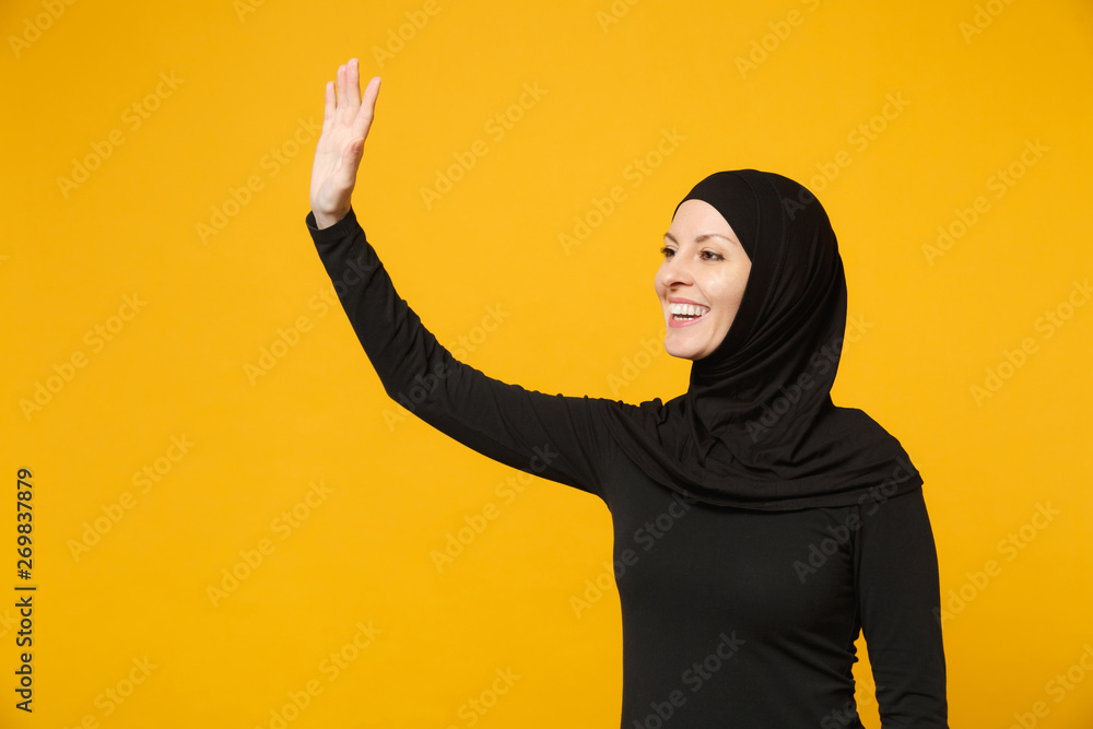 Young arabian muslim woman in hijab black clothes waving and greeting with hand as notices someone isolated on yellow wall background. People religious Islam lifestyle concept. Mock up copy space. Young arabian muslim woman in hijab black clothes waving and greeting with hand as notices someone isolated on yellow wall background. People religious Islam lifestyle concept. Mock up copy space.
