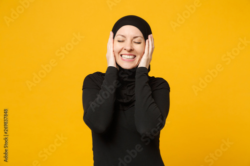 Smiling confident beautiful young arabian muslim woman in hijab black clothes posing isolated on yellow wall background, studio portrait. People religious Islam lifestyle concept. Mock up copy space. © ViDi Studio