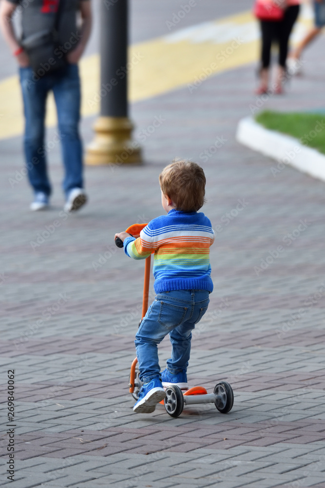 Fototapeta premium Boy child in blue jeans and sneakers riding a scooter. back view