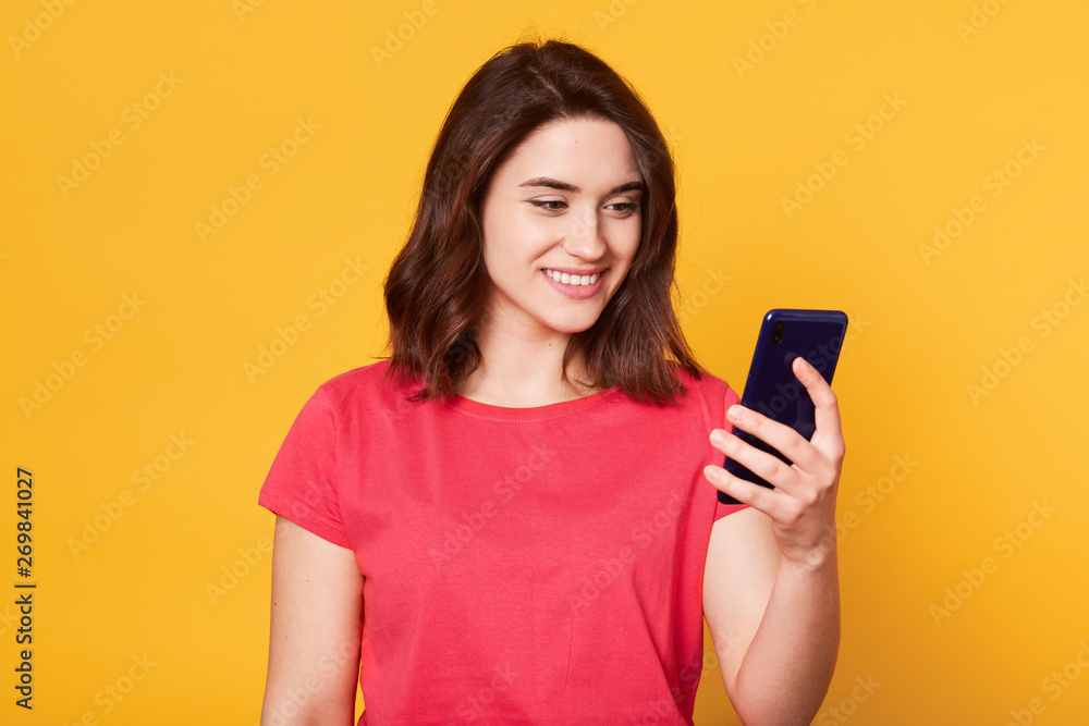 Studio shot of young good looking European woman with dark hair isolated on yellow background, holding smart phone in hand, looking at screen smiling while checking social network or reading message.