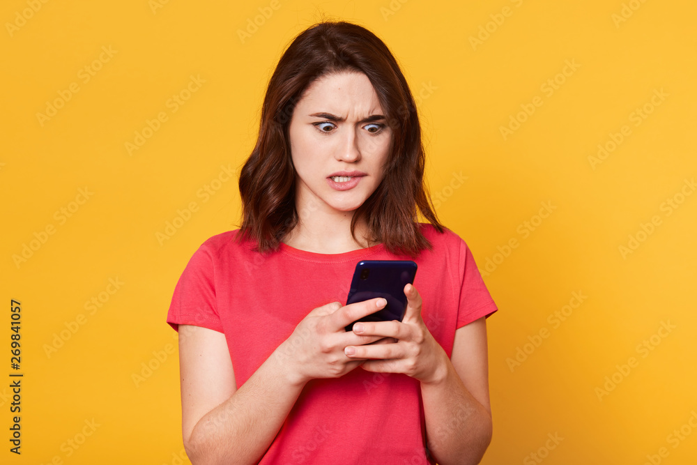 Image of scared young brunette female biting her lip, feels fear, holding modern smart phone, wears casually, reads something astonishing in internet, isolated over yellow studio background. Image of scared young brunette female biting her lip, feels fear, holding modern smart phone, wears casually, reads something astonishing in internet, isolated over yellow studio background.
