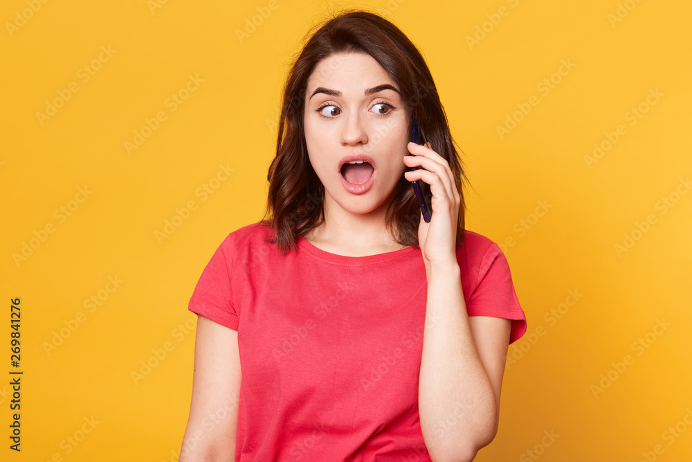 Close up portrait of amazed beautiful girl in casual style, has dark hair, standing and speaking on mobile smart phone and looking with shocked face and open mouth isolated over yellow background. Close up portrait of amazed beautiful girl in casual style, has dark hair, standing and speaking on mobile smart phone and looking with shocked face and open mouth isolated over yellow background.