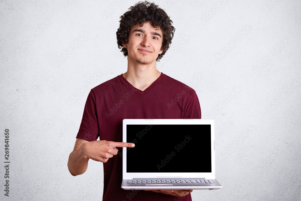 Shot of smiling handsome man wearing casual maroon t shirt standing and pointing finger on blank laptop screen over white studio background, male looking at camera with plasant facial expression. Shot of smiling handsome man wearing casual maroon t shirt standing and pointing finger on blank laptop screen over white studio background, male looking at camera with plasant facial expression.
