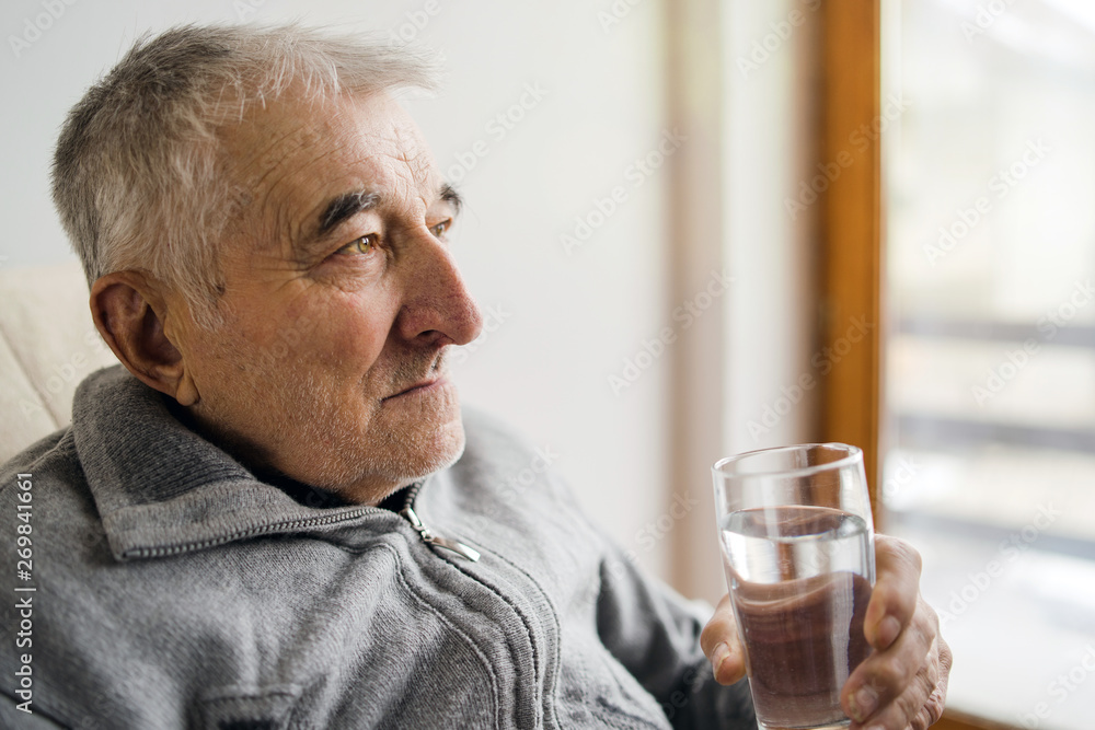 Senior man sitting holding glass of water looking sad by the window at the retirement nursing home Senior man sitting holding glass of water looking sad by the window at the retirement nursing home
