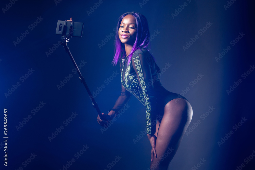 Selfie girl. Black afro model takes a photo with a selfie stick. Girl in a black bodysuit posing. Selfshot. Portrait with a selfie stick. Photo for social networks. Egomania. Fashion trend.