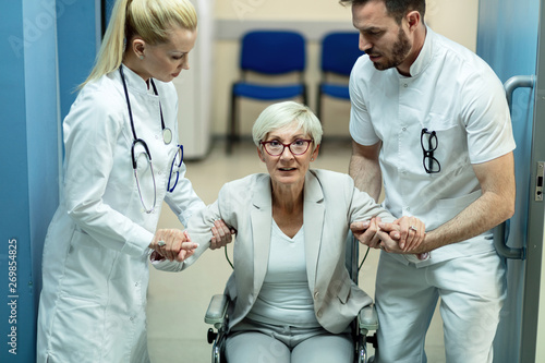 Photos Two doctors assisting mature patient to get up from the wheelchair in the hospital