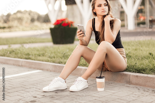 Beautiful young woman using phone and drink coffee sitting on the curb in the street.