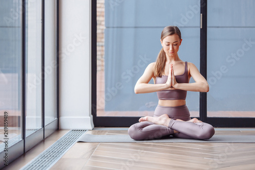 Relaxed young girl practicing yoga at home sitting near the large windows of a country house. Concept of meditation and relaxation. Ardha Padmasana exercise, meditating in Lotus pose with namaste,