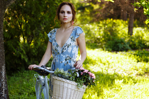 portrait of a beautiful girl in the forest, holding a bike with a basket of flowers, behind the rays of the sun, a blue flowered dress, summer walk © Skripnik Olga