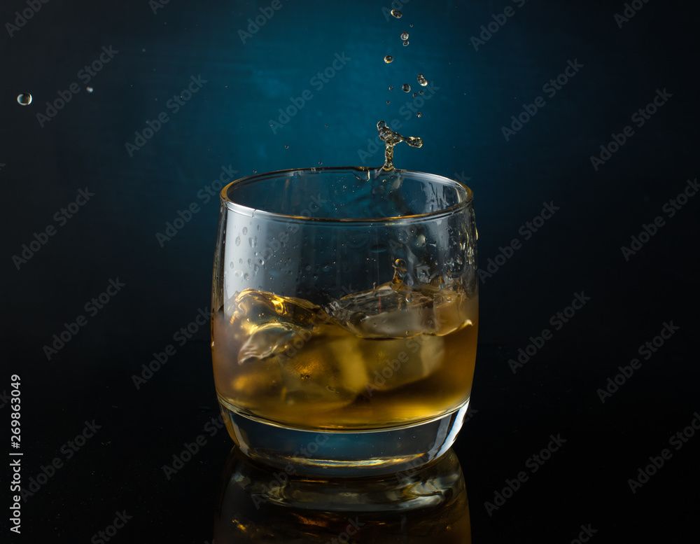 A glass of whiskey with ice on a dark color photo with splashes from falling ice A glass of whiskey with ice on a dark color photo with splashes from falling ice