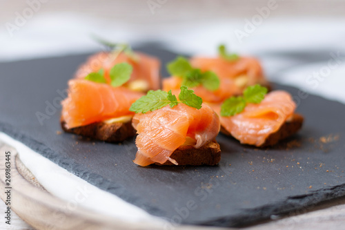 Smoked salmon on toast with butter with mint leaf on black stone and old wooden board - image