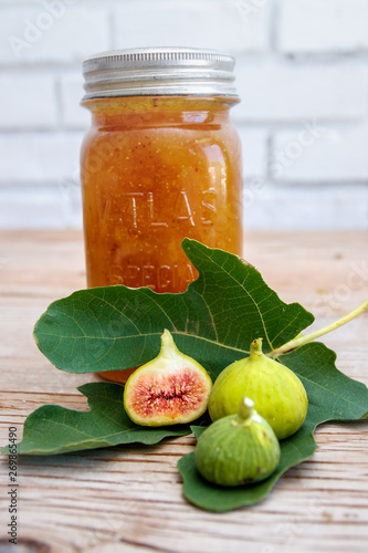 Antique glass preserve bottle with fig preserve together with a fresh green fig leaf and halved ripe figs on a raw wood surface. 