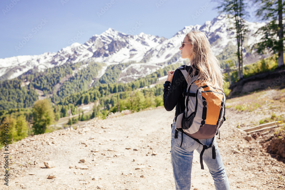 Naklejka premium Rear view of a beautiful young independent girl blonde traveler travels with a backpack in the mountains. Concept of desire for solitude and freedom.