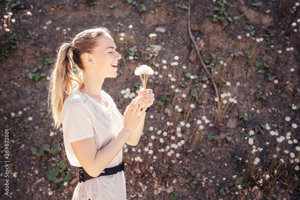 Happy young woman rejoices in the beauty of nature by blowing a dandelion in the wind on a sunny day. The concept of vacation alone in the summer and avoiding the city rush Happy young woman rejoices in the beauty of nature by blowing a dandelion in the wind on a sunny day. The concept of vacation alone in the summer and avoiding the city rush