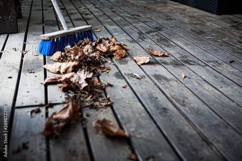 Focus on a blue broom with dry, brown autumn leaves on a dirty, dusty wooden deck. Foreground is out of focus.
