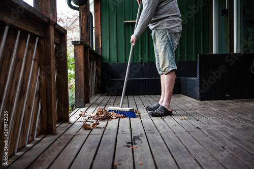 A pile of old and dry autumn leaves on a wooden deck. A man is using a blue broom to sweep and spring clean the porch, preparing it for the summer. 