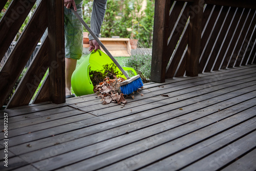 A green plastic basket is used to collect a pile of old and dry leaves from a porch. A man is using a broom to sweep the waste into the basket. Spring cleaning.