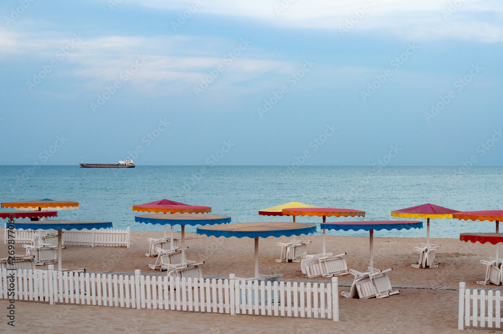 Obraz premium Desert seascape with white deck chairs under colorful beach umbrellas on dusky sky background. White wood fence on beach sand. Blurred silhouette of cargo ship on skyline. Vacation at Black sea coast 