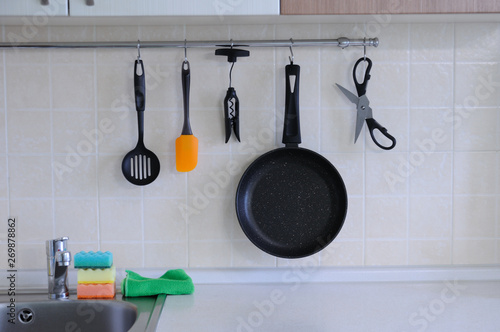 Non-stick frying pan, scissors for food, a spatula for turning steaks, a spoon, a corkscrew against a tiled wall. The interior of the kitchen.