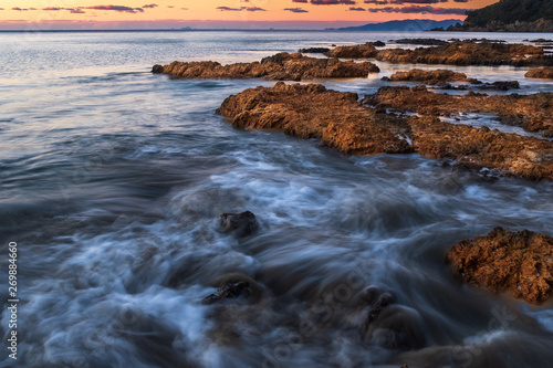 Waves crashing over rocks on the jagged coastline of Waiheke Island, New Zealand, taken on a long exposure to smooth out the water and create beautiful smooth effect, against a sunset nobody in the