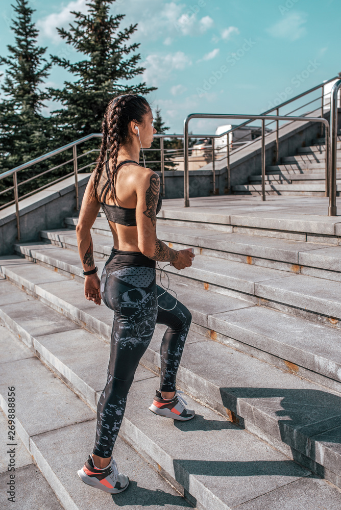 Athlete woman walks sports hot workout summer city. Headphones phone, online application music social networks. Concept of fitness in fresh air, an active lifestyle. Tattoos on tanned figure.