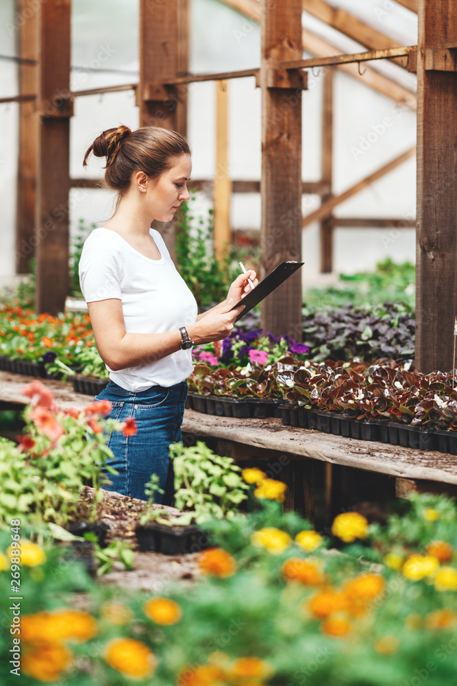 Young woman agronomist inspecting plants in greenhouse. Female gardener working in garden with flowers Young woman agronomist inspecting plants in greenhouse. Female gardener working in garden with flowers