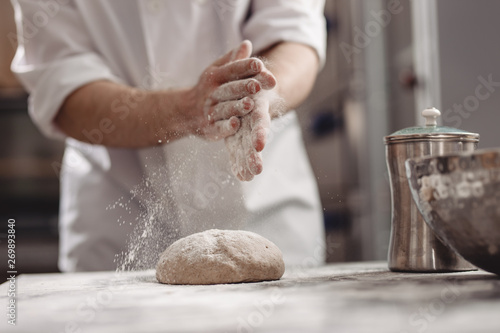 Baker adds flour to dough on the table in the bakery
