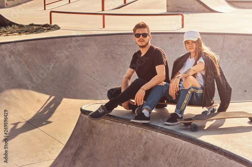 Cool trendy students are sitting at sunny skatepark with their longboards. There are other skatebordists at background.