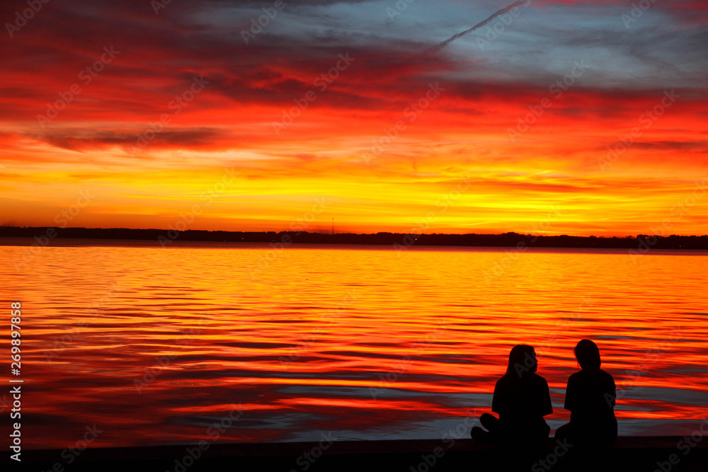 Two girls sitting with a bright orange sunset on the lake