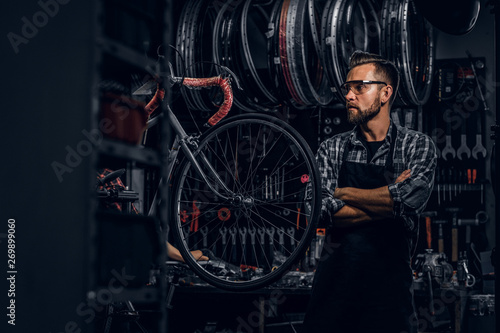 Smiling bearded man in glasses is standing near fixed bicycle at his own workshop.