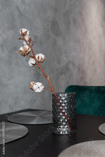 Dried cotton branch in stylish ceramic vase stands on black kitchen table. Interesting modern interior details. Grey wall background.