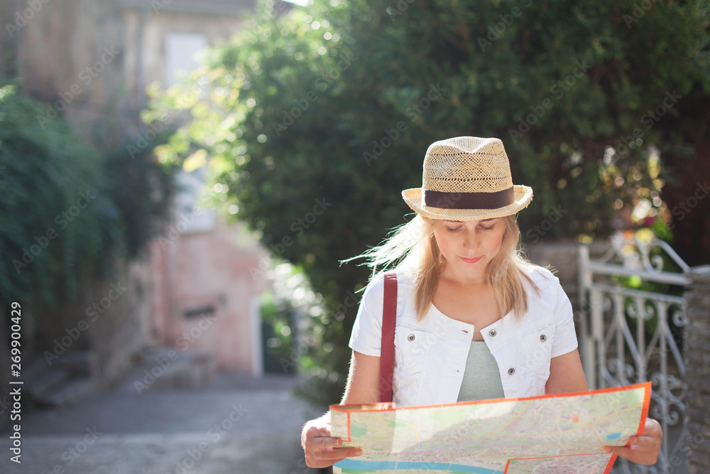 Fototapeta premium Woman traveler in hat looks at city map at town street in Europe. Girl tourist is searching apartment, housing. Concept of student travel, summer vacation, solo female tourism, adventure, journey.