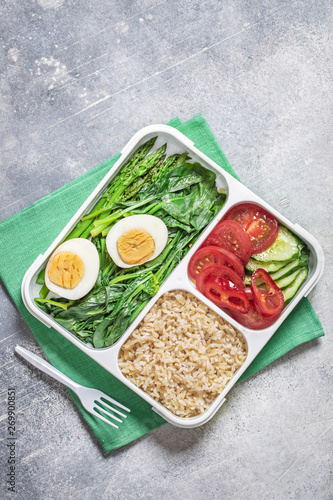 Brown rice, stewed spinach and asparagus with vegetables and egg, healthy lunch box on gray concrete background, top view 