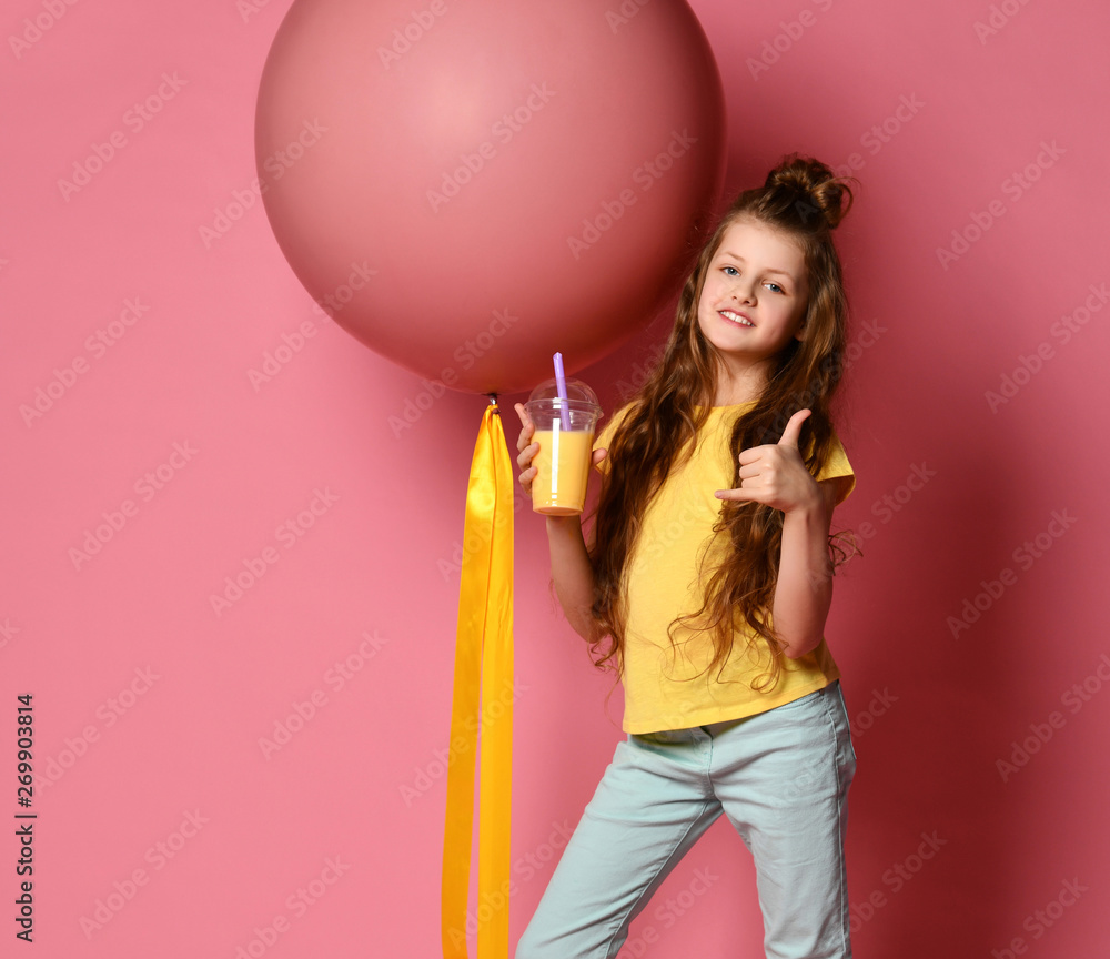 Pleased and happy young lady, teen girl in jeans and yellow t-shirt with glass of yellow juice stands at big pink balloon Pleased and happy young lady, teen girl in jeans and yellow t-shirt with glass of yellow juice stands at big pink balloon