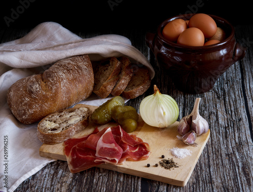Traditional snacks with slices of ham and vegetables, pickled cucumbers, onion, garlic, pepper, salt, bread and butter, eggs in a clay pot on a dark rustic wooden table