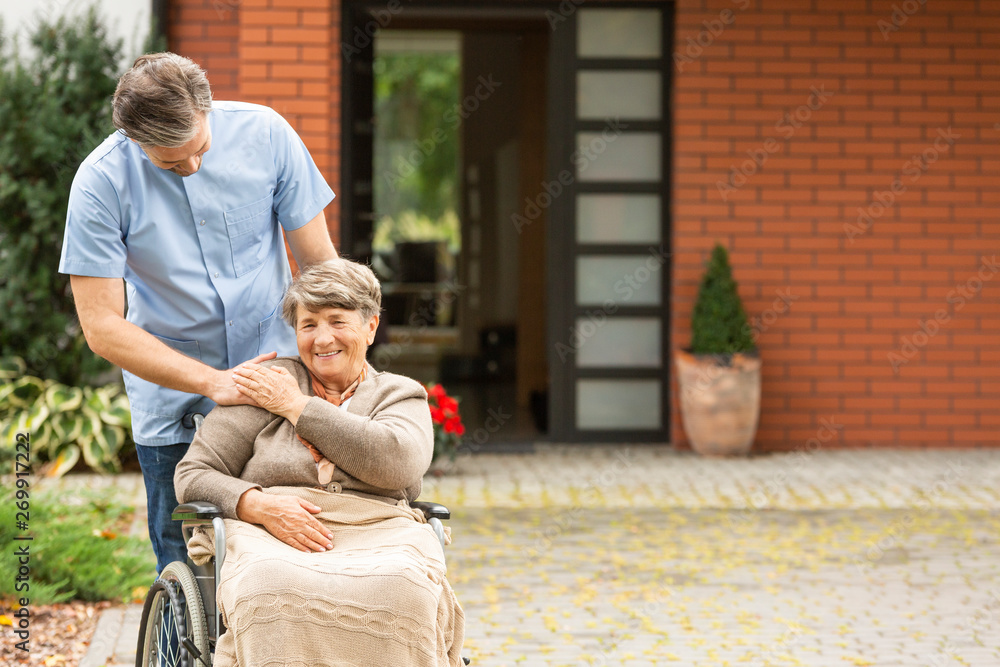 Caregiver helping smiling disabled senior woman in the wheelchair in front of house - obrazy, fototapety, plakaty 