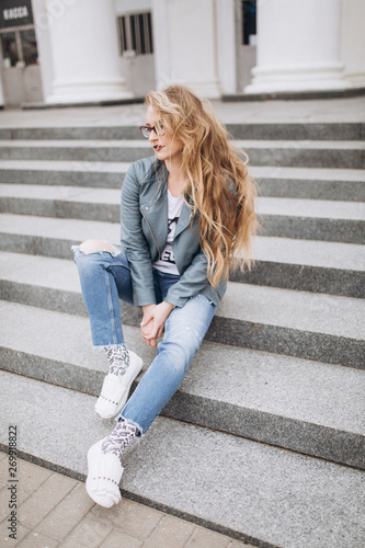 Close up portrait of young pretty caucasain woman in glasses with long blondie hair sitting on stairs outdoors dreaming. Happiness, freedom, beauty concept