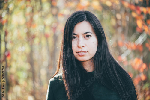 Female beauty portrait surrounded by vivid foliage. Dreamy beautiful girl with long natural black hair on autumn background with colorful leaves in bokeh. Inspired girl enjoys nature in autumn forest.